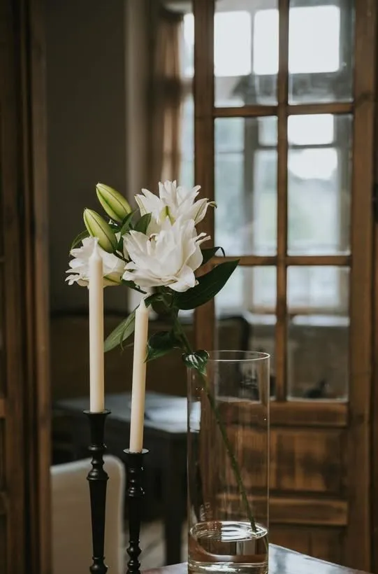 Baltas lilijas augstā stikla vāzē blakus divām svecēm melnos svečturos uz koka galda